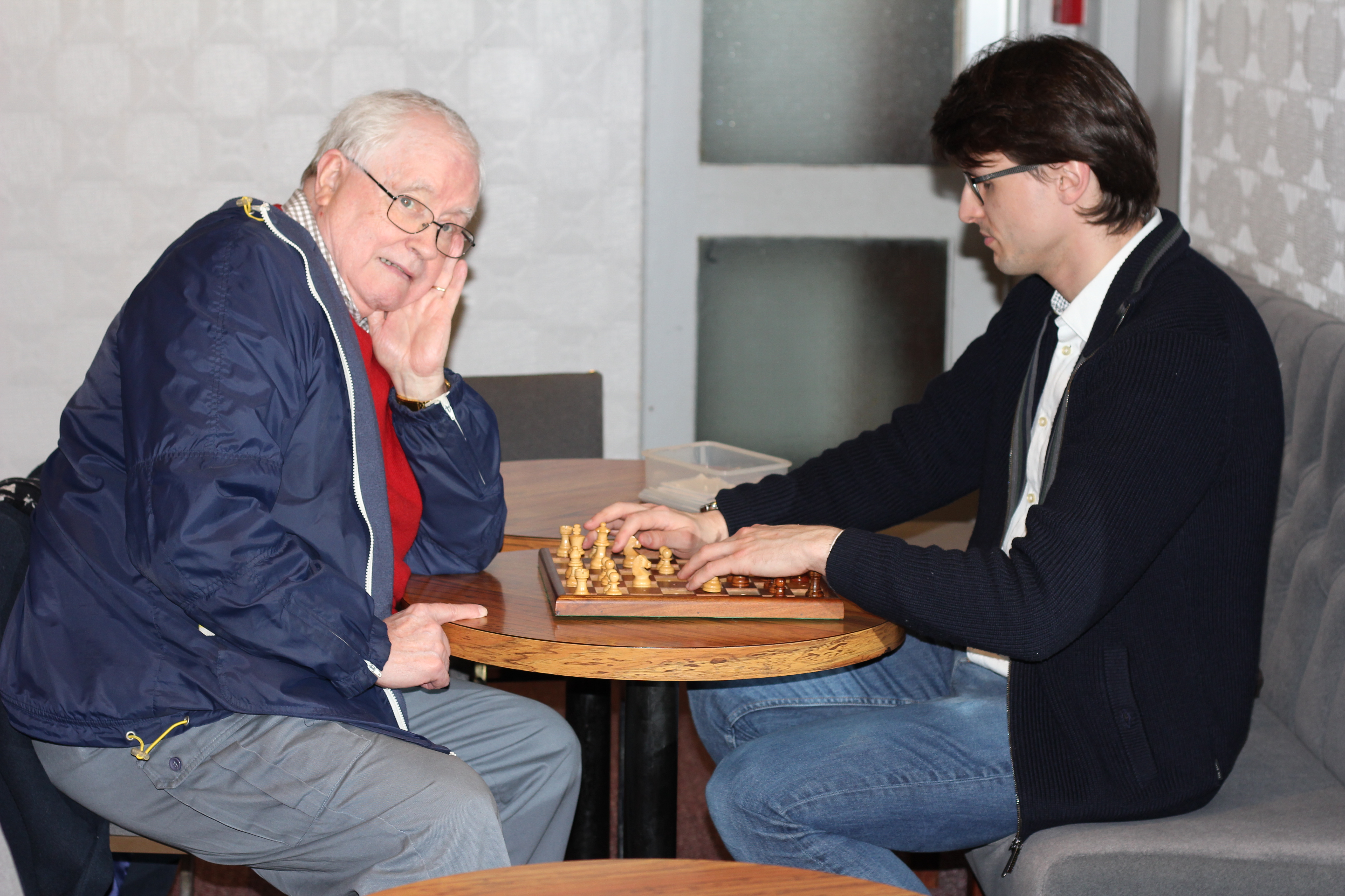 Two players sat at a braille chess board. One of the players is blind, and is feeling the pieces before his turn.